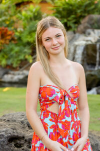 Emily Matejka, Legal Assistant smiling outdoors in a bright red and blue floral dress, with greenery and rocks behind her.