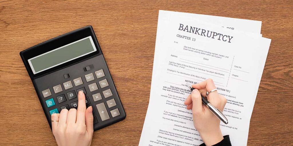 Hands using a calculator and signing official Chapter 13 bankruptcy forms, illustrating the process when a tenant files for bankruptcy.