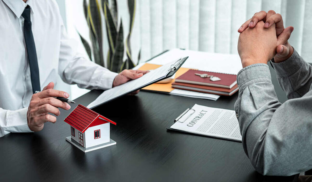 A landlord holding documents and pointing at a small house model while sitting across from a tenant during a lease discussion.