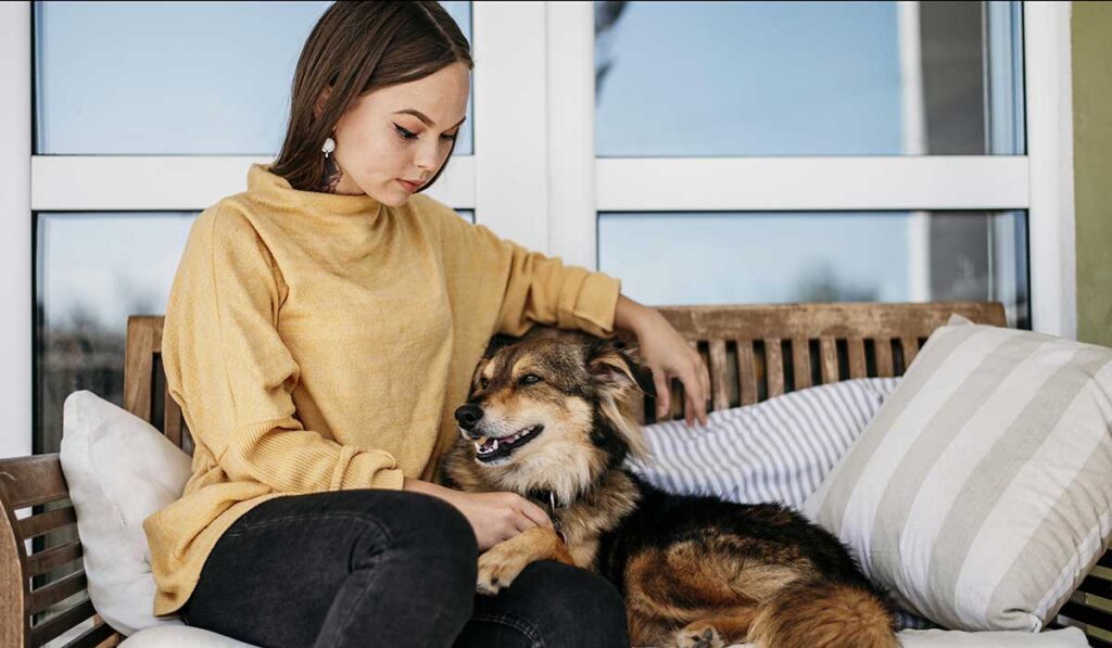 A woman gently rests her hand on her dog while sitting together on a wooden bench near large glass windows.