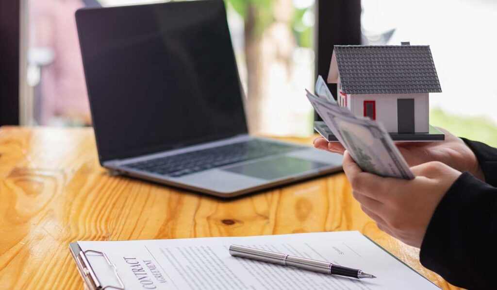 : A person holds cash beside a miniature house and lease agreement documents during a rental property payment transaction.